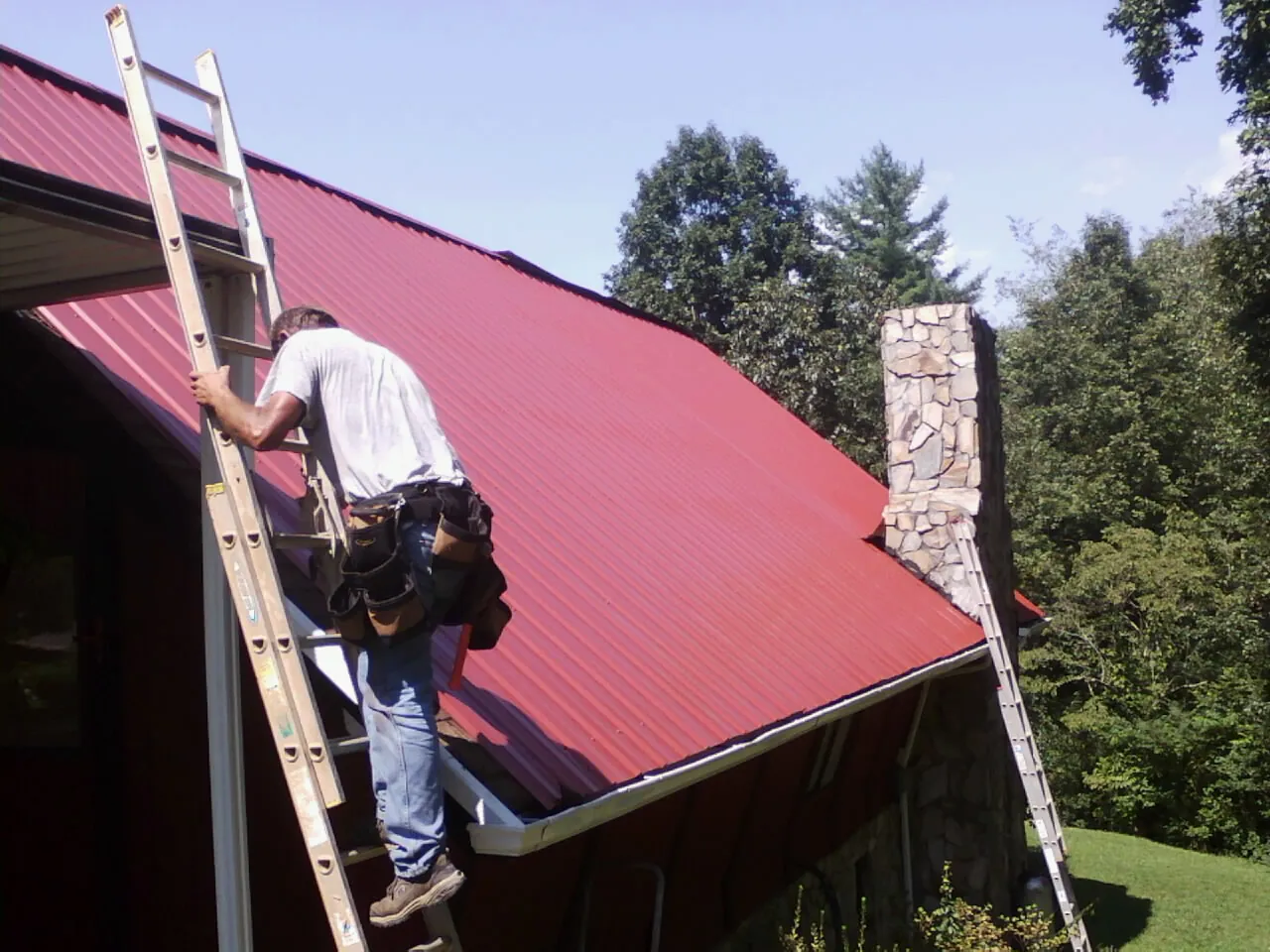 Roofer on a ladder beside a red metal roof and stone chimney — Soffit & Fascia Repair by Roofing Brothers in Olalla
